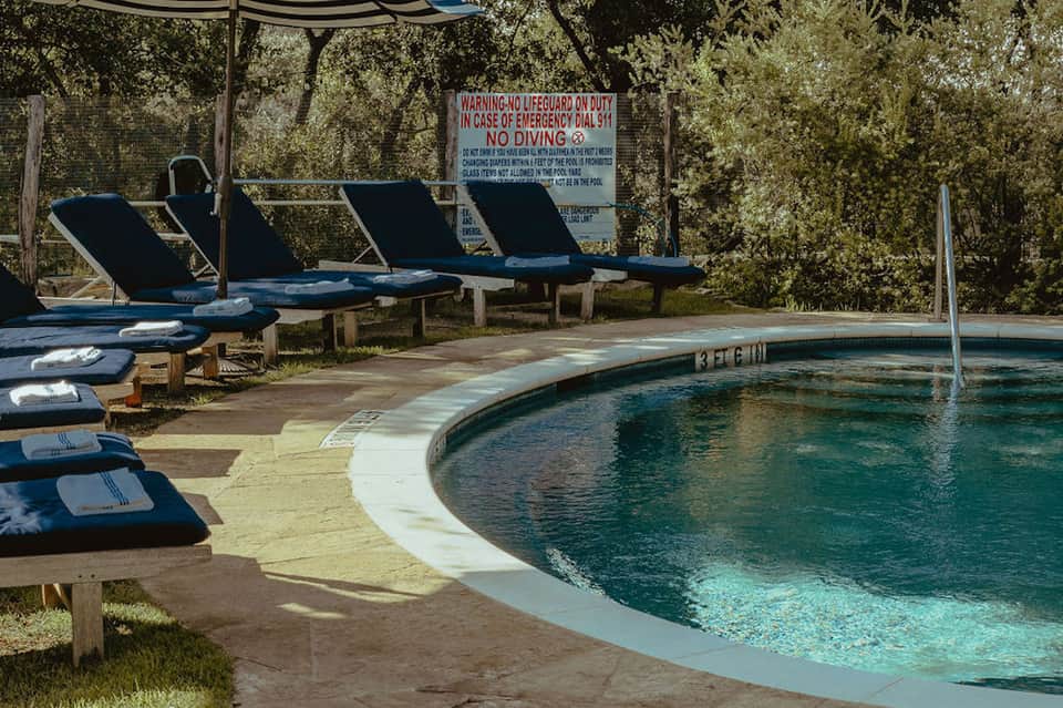 Small resort pool surrounded by lounge chairs under pergola with warning sign and natural landscaping