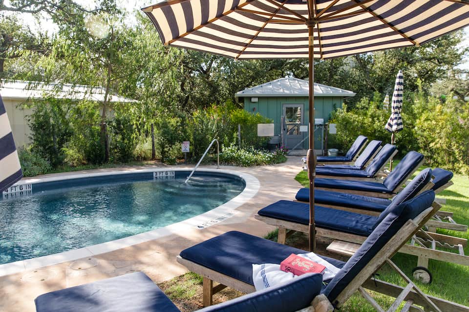 Outdoor hot tub surrounded by lounge chairs with blue umbrellas and lush green landscaping