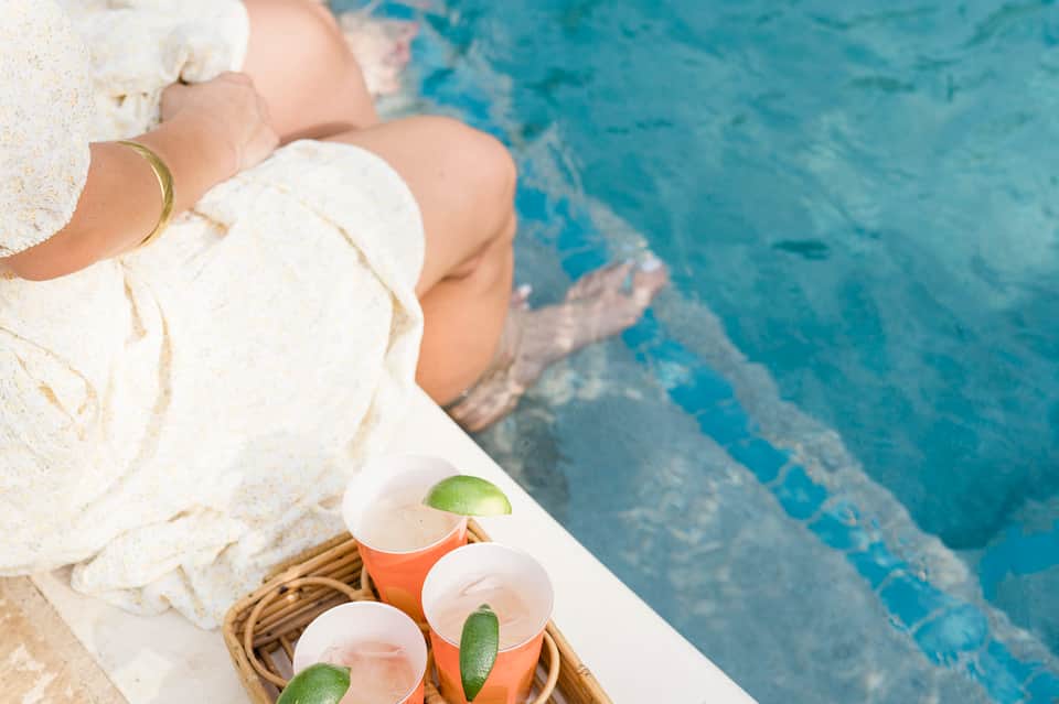 Close-up of person in white towel holding tropical cocktails poolside with turquoise water