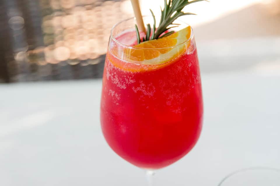 Tropical cocktail with red liquid, citrus garnish, and rosemary sprig on white surface