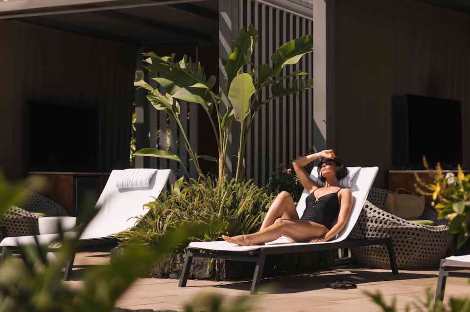 Woman relaxing on white lounge chair in modern courtyard with tropical plants and black pergola