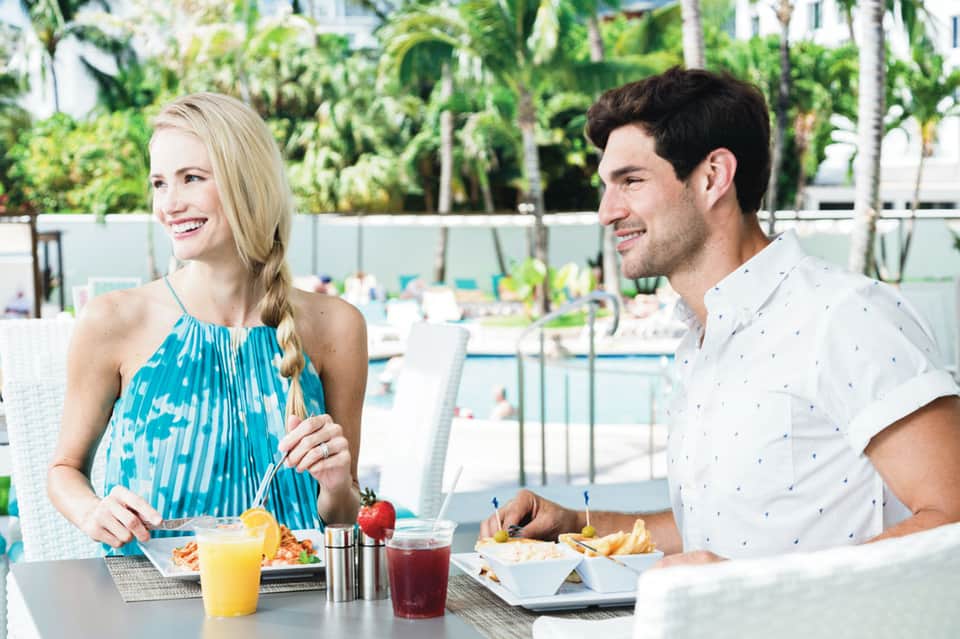 Couple dining poolside with salads and beverages, tropical garden backdrop visible