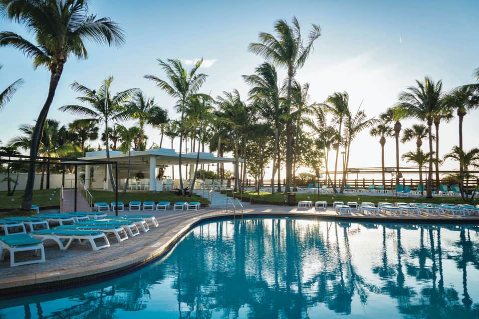 Curved resort pool surrounded by palm trees with blue lounge chairs and a poolside pavilion at dawn