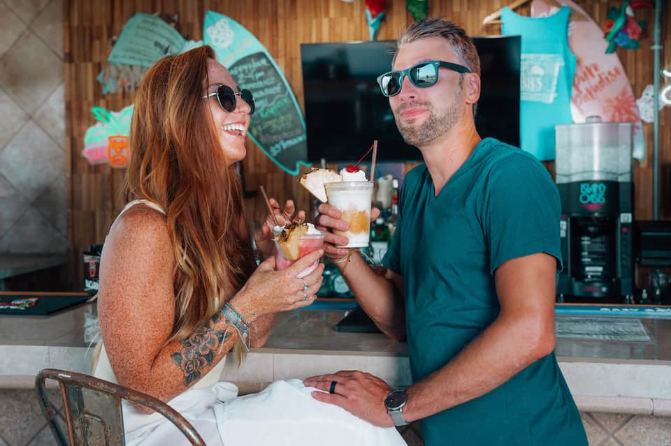 Couple in casual beach attire enjoying tropical cocktails at tiki bar with surfboards and colorful decor