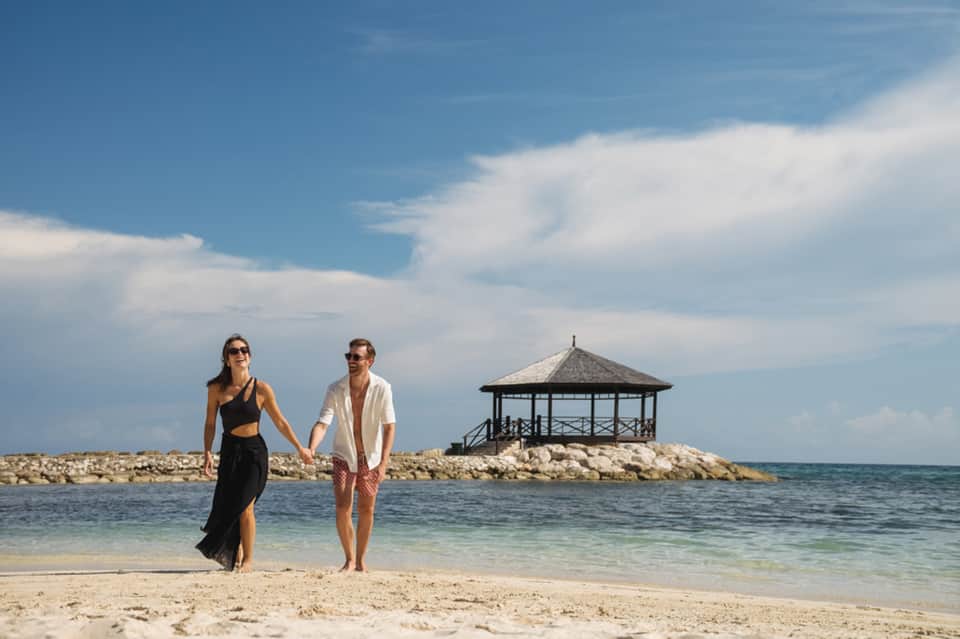 Couple walking on beach holding hands with gazebo pavilion on rocky outcrop in background