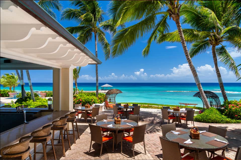 Beachfront restaurant with palm trees, ocean views, dining tables, and modern white pergola