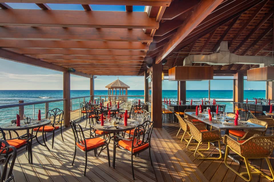 Beachfront dining area under wooden pergola with ocean views and orange chair cushions
