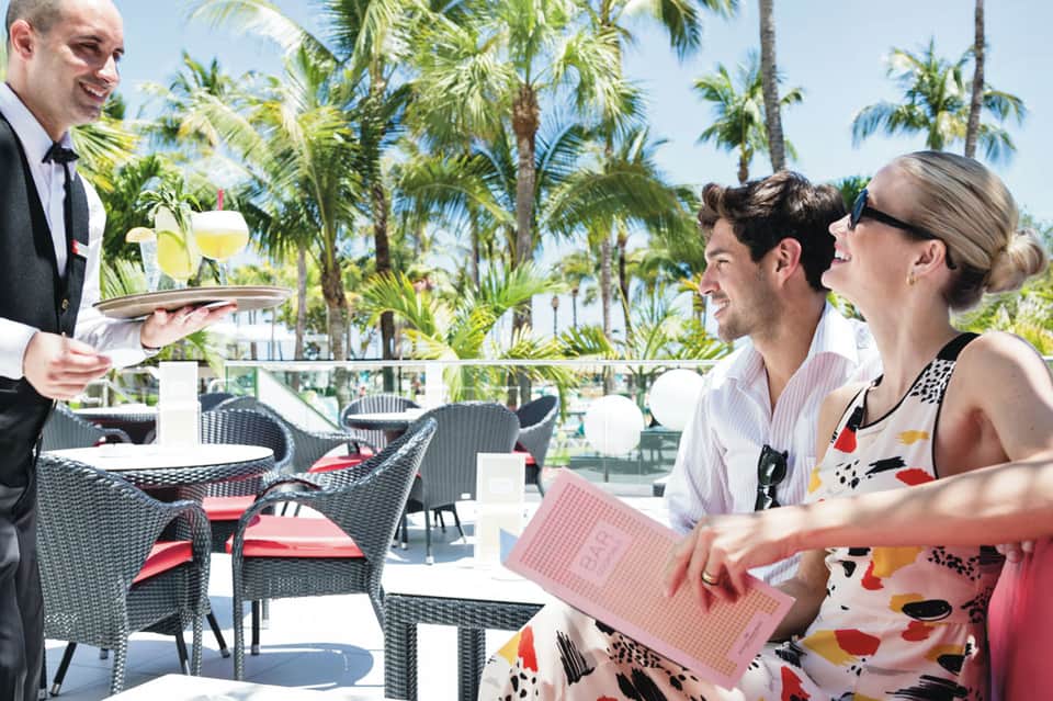 Waiter serving tropical drinks to couple at beachside resort terrace with palm trees