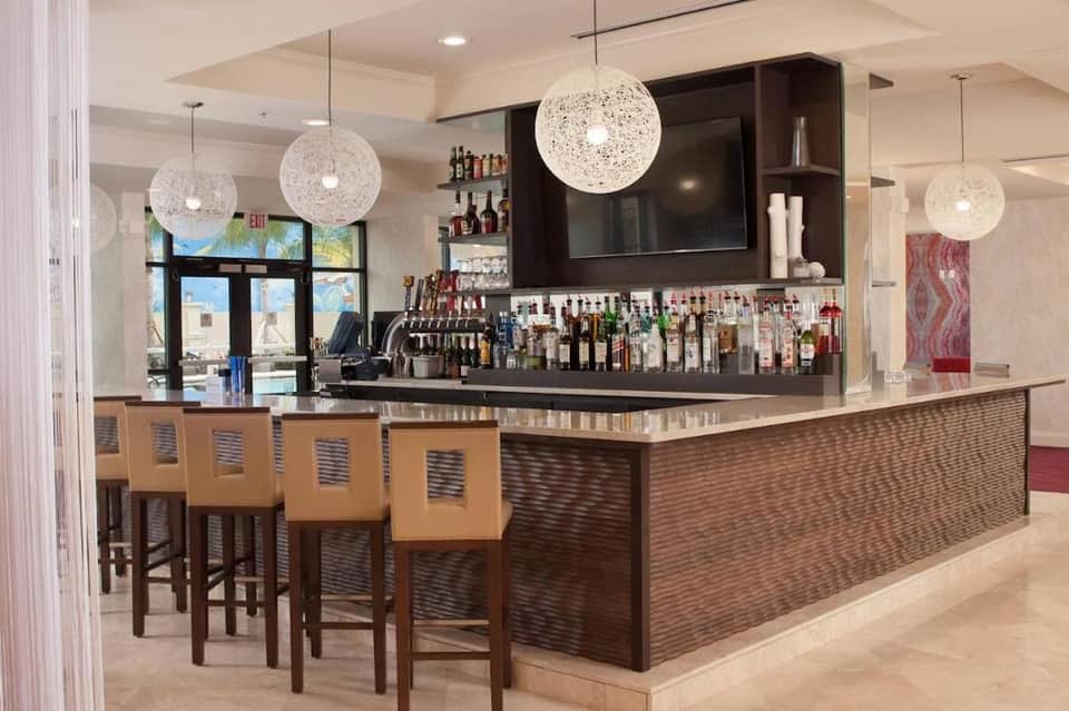 Modern hotel bar with white countertop, dark shelving, woven pendant lights, and wooden bar stools with yellow cushions
