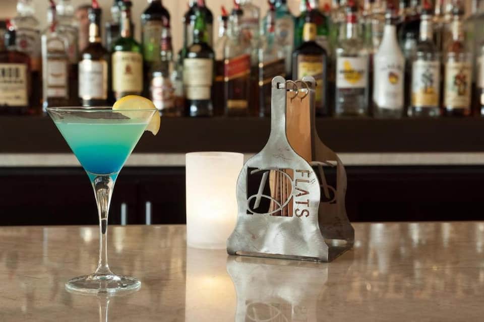 Turquoise cocktail with lemon slice on bar counter with full liquor bottle collection blurred in background