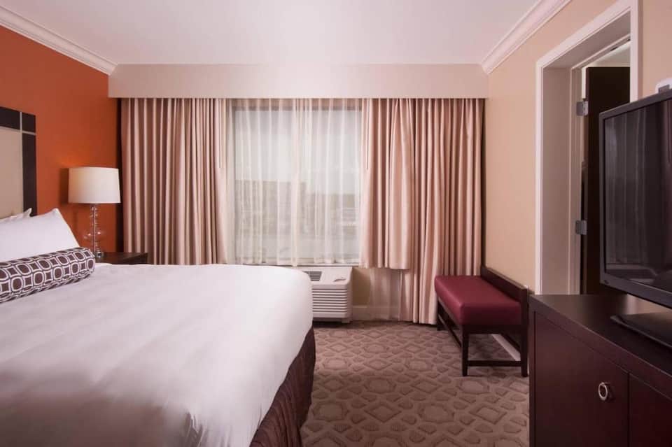 Hotel bedroom with queen bed, orange accent wall, beige curtains, and modern furnishings