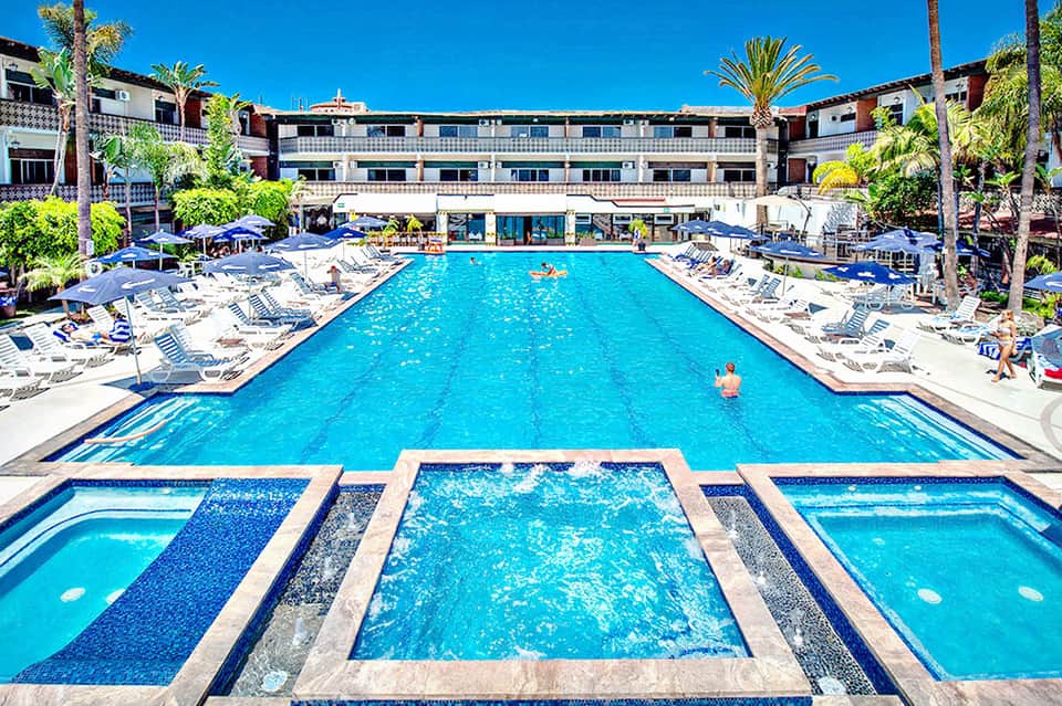 Resort courtyard with large rectangular pool surrounded by lounge chairs, palm trees, and multi-story hotel buildings under clear blue sky