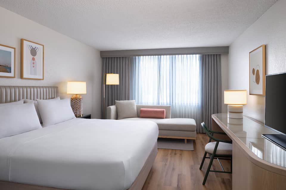 Contemporary hotel bedroom with queen bed, seating area, and large window with curtains