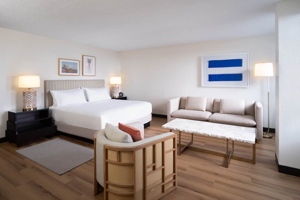 Hotel suite with king bed, seating area, striped blue artwork, and modern coastal-inspired décor