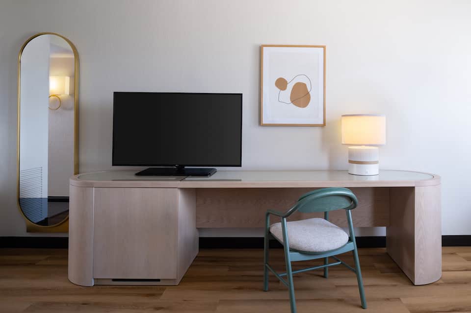 Modern hotel room desk with curved wood design, TV, mirror, and chair with city view