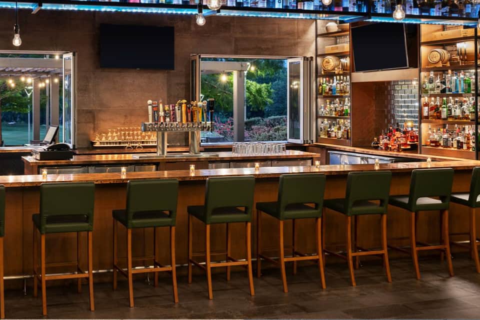 Modern bar interior with wooden counter, green bar stools, illuminated shelving with liquor bottles, and glass exterior view