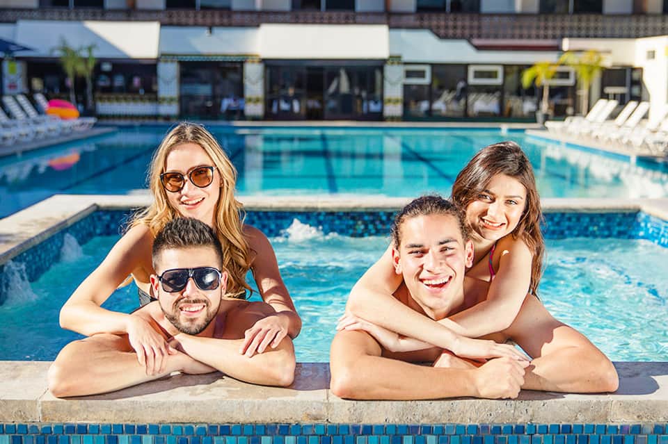 Four young adults smiling at the edge of a resort swimming pool with lounge chairs and buildings in the background