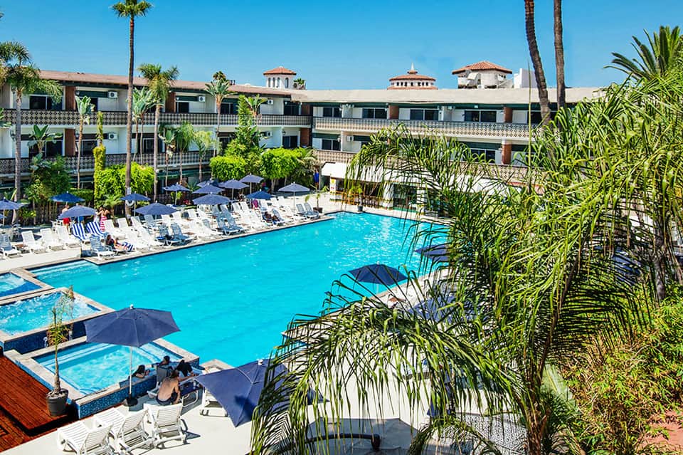 Large resort pool with turquoise water, white loungers, blue umbrellas, Spanish-style buildings, and palm trees