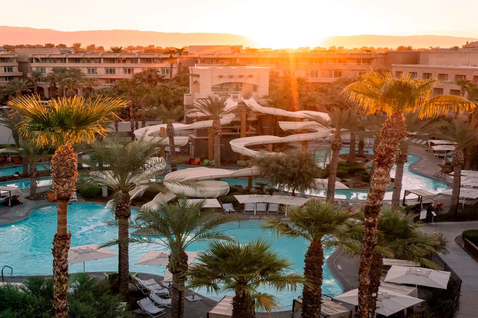 Aerial view of resort pools with turquoise water, palm trees, and buildings at golden sunset