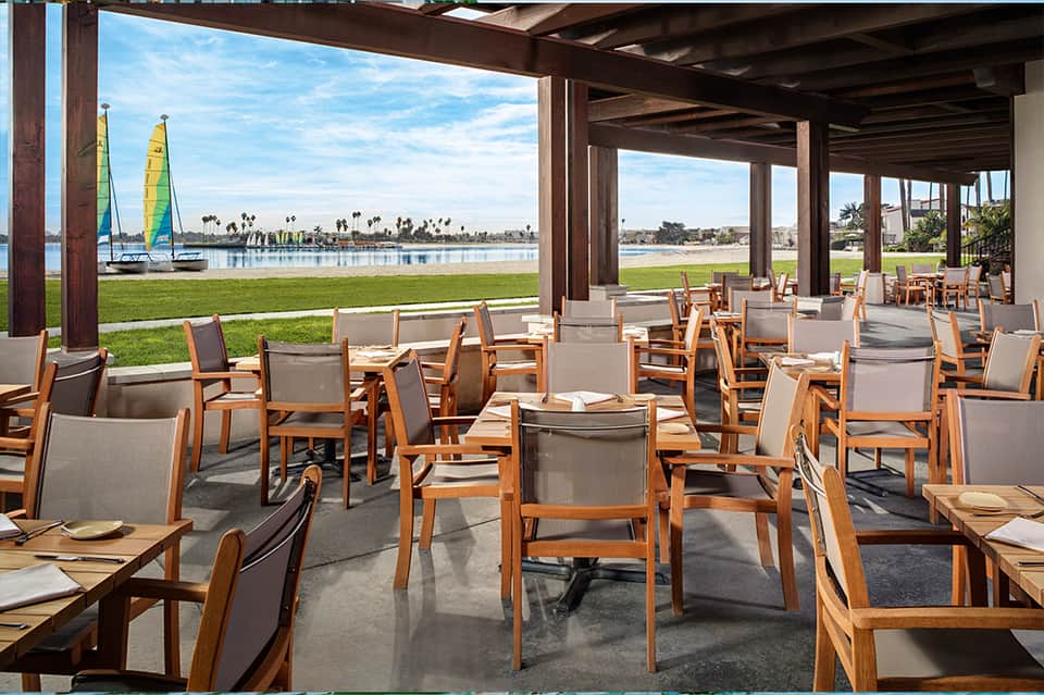 Waterfront dining pavilion with wooden chairs, water views, sailboats, and palm trees