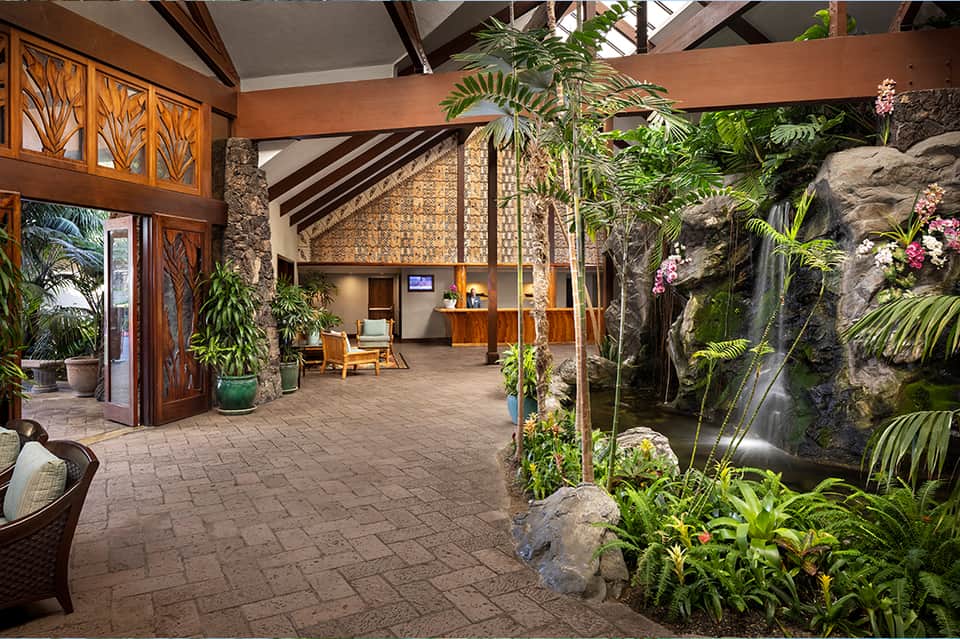 Tropical resort atrium with lush plants, stone waterfall, wooden beams, and natural lighting