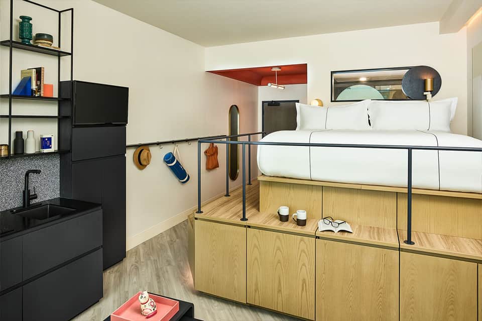 Modern hotel room with loft bed, wooden storage, kitchenette, and metal railing