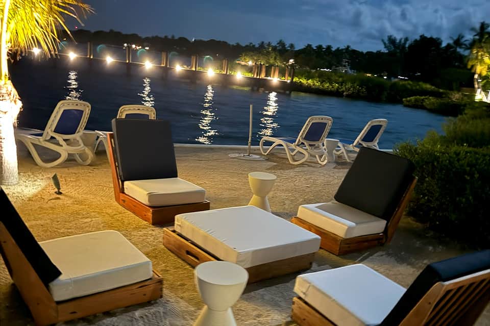 Illuminated waterfront lounge area at night with wooden deck chairs and palm trees