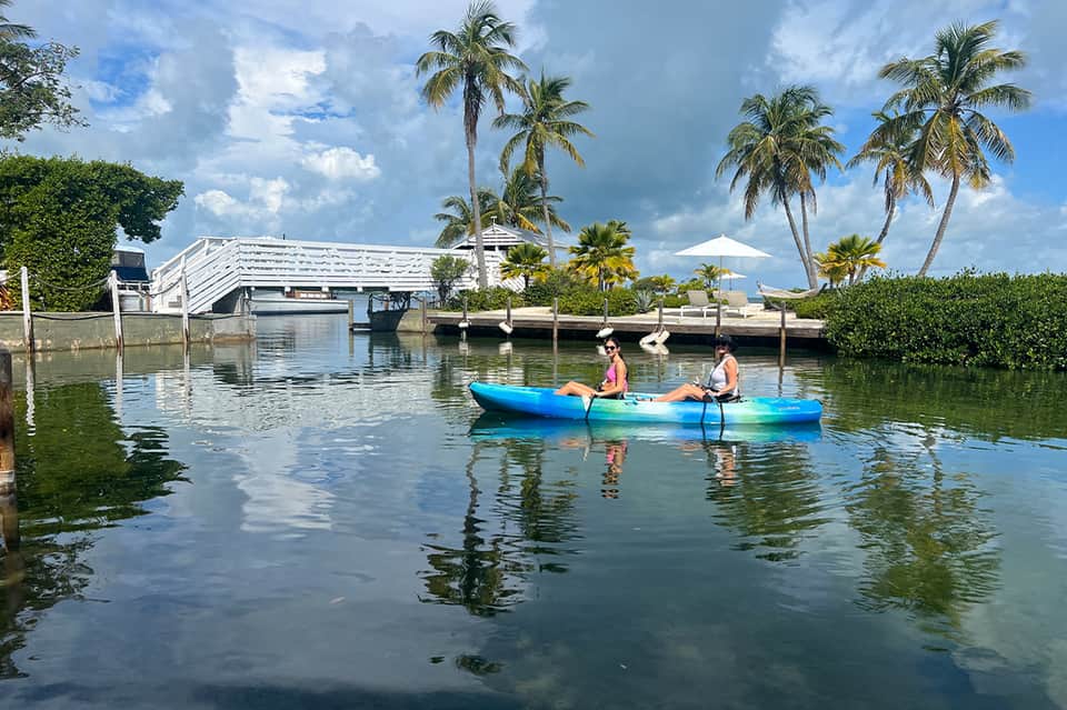 Two people kayaking on calm waterway lined with palm trees near modern waterfront building