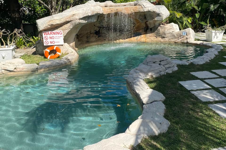 Lagoon-style resort pool with stone waterfall, stepping stones, tropical landscaping, and safety signage