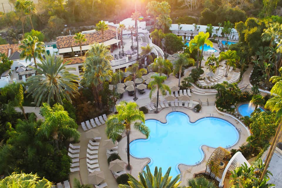 Aerial view of resort with curved pool, cabanas, palm trees, and surrounding landscaping at sunset
