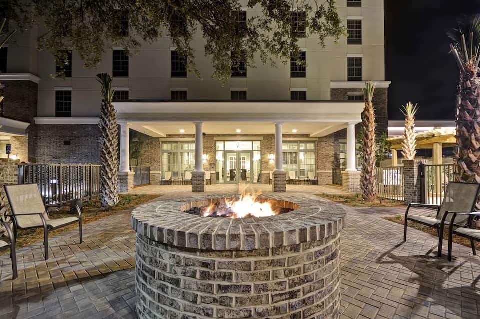 Outdoor courtyard at night with firepit surrounded by seating, illuminated building entrance, and palm trees
