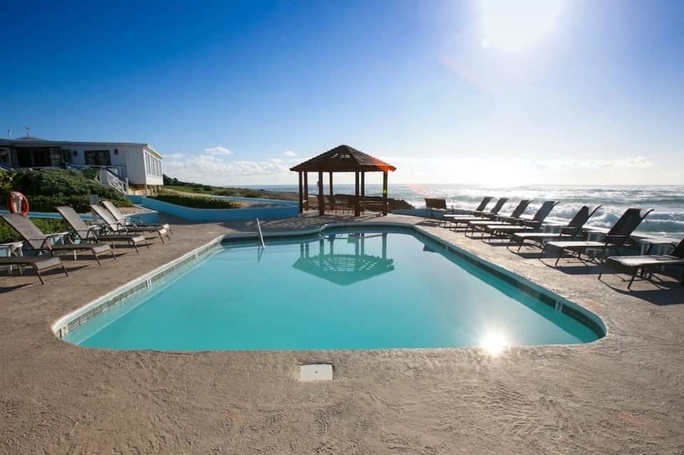Beachfront resort pool with turquoise water, lounge chairs, gazebo, and ocean view under clear blue sky