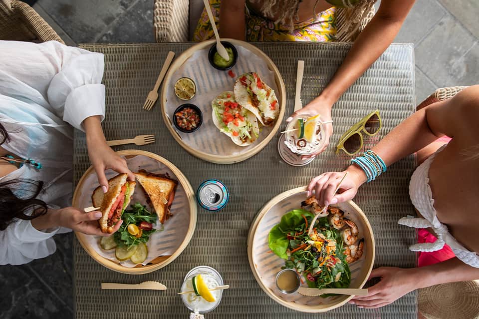 Overhead view of people dining outdoors with tacos, sandwiches, and salads on wooden plates