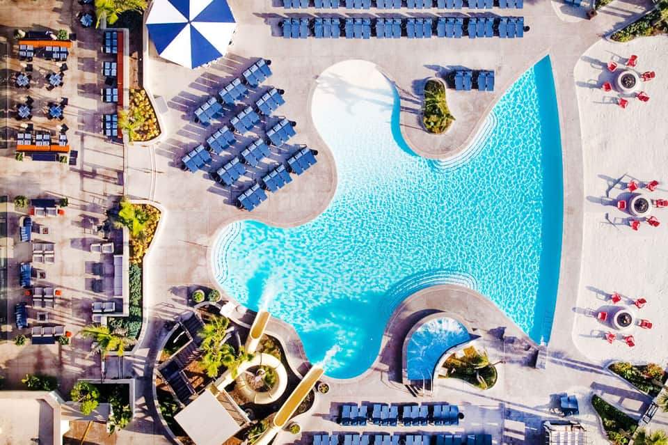 Aerial view of resort pool with blue loungers, umbrellas, and surrounding buildings