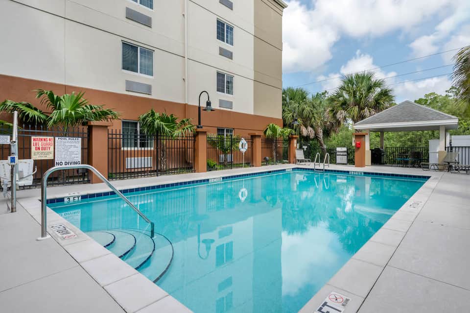 Rectangular hotel pool with ladder, white pergola, palm trees, and orange building exterior