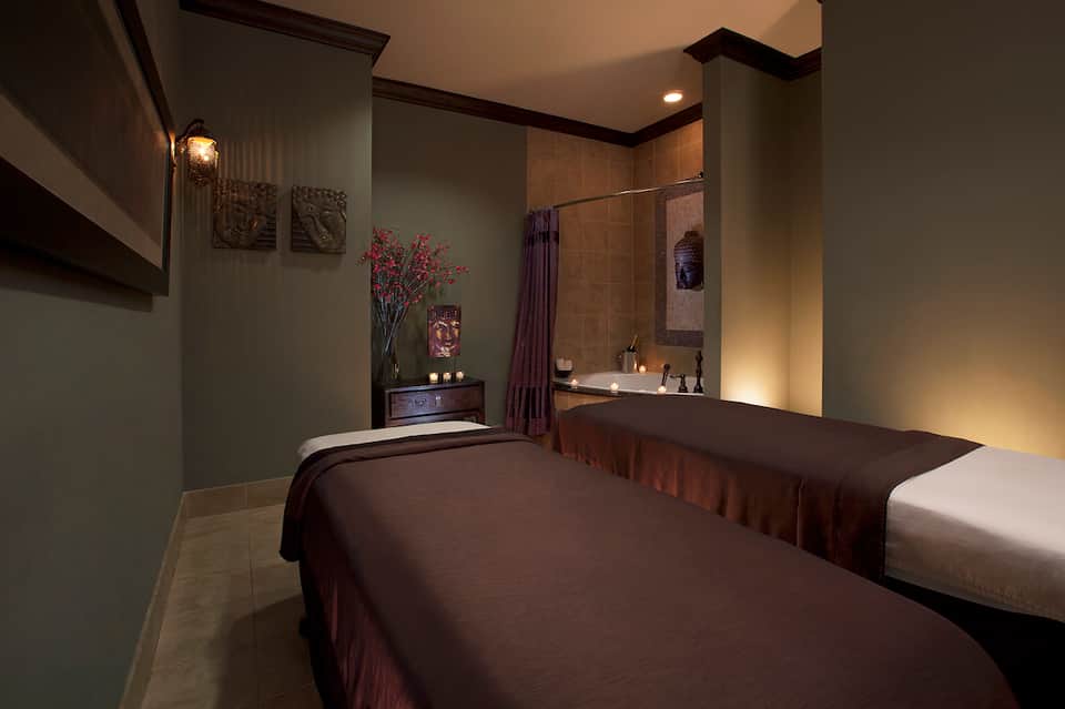 Spa treatment room with two massage tables, green walls, decorative flowers, and ambient lighting