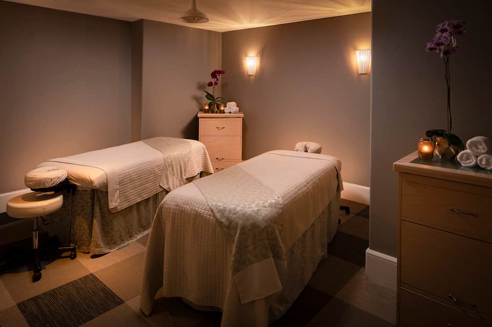 Spa treatment room with two massage tables draped in cream linens and soft ambient lighting