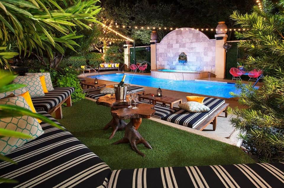 Nighttime resort pool area with striped loungers, tropical plants, illuminated water features, and fire pit