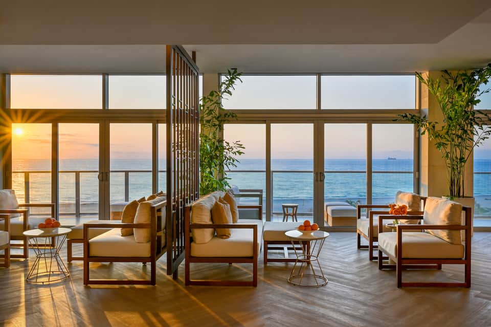 Luxury oceanfront lounge at sunset with modern furniture, floor-to-ceiling windows, and wooden accents