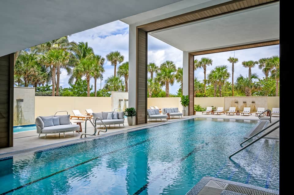 Indoor swimming pool with floor-to-ceiling windows, palm tree views, and lounge seating
