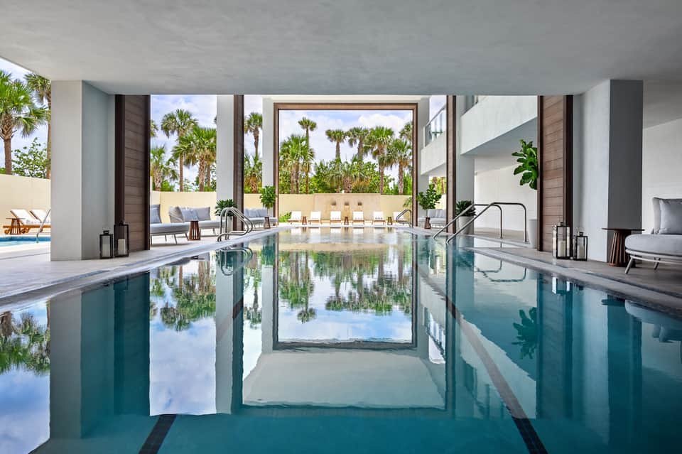Indoor lap pool with glass doors opening to outdoor patio and palm trees reflected in water