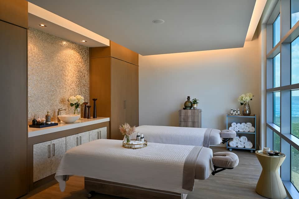 Luxurious spa treatment room with massage beds, vanity, towel storage, and ocean views