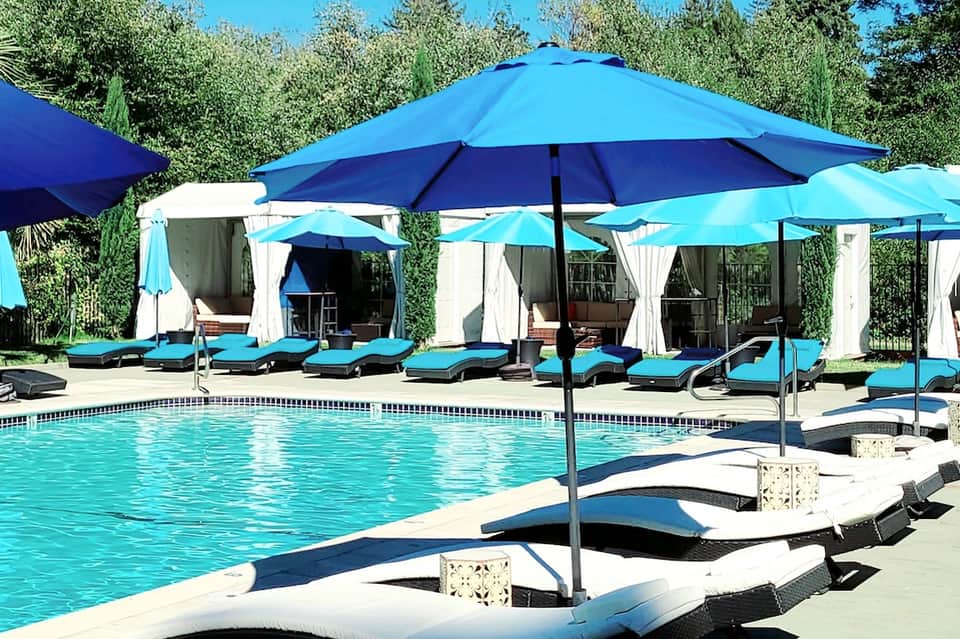 Resort pool area with turquoise loungers, large blue umbrellas, and white cabanas surrounded by trees