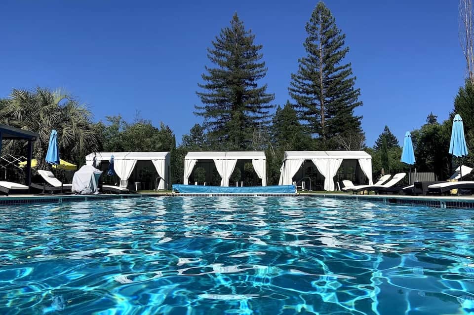 Resort outdoor pool with crystal-clear water, white cabanas, blue umbrellas, and tall pine trees in background