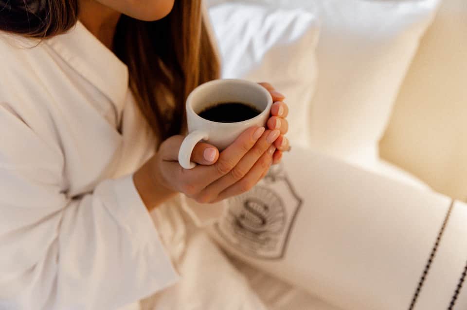 Woman in white clothing holding white cup of black coffee indoors on light neutral background