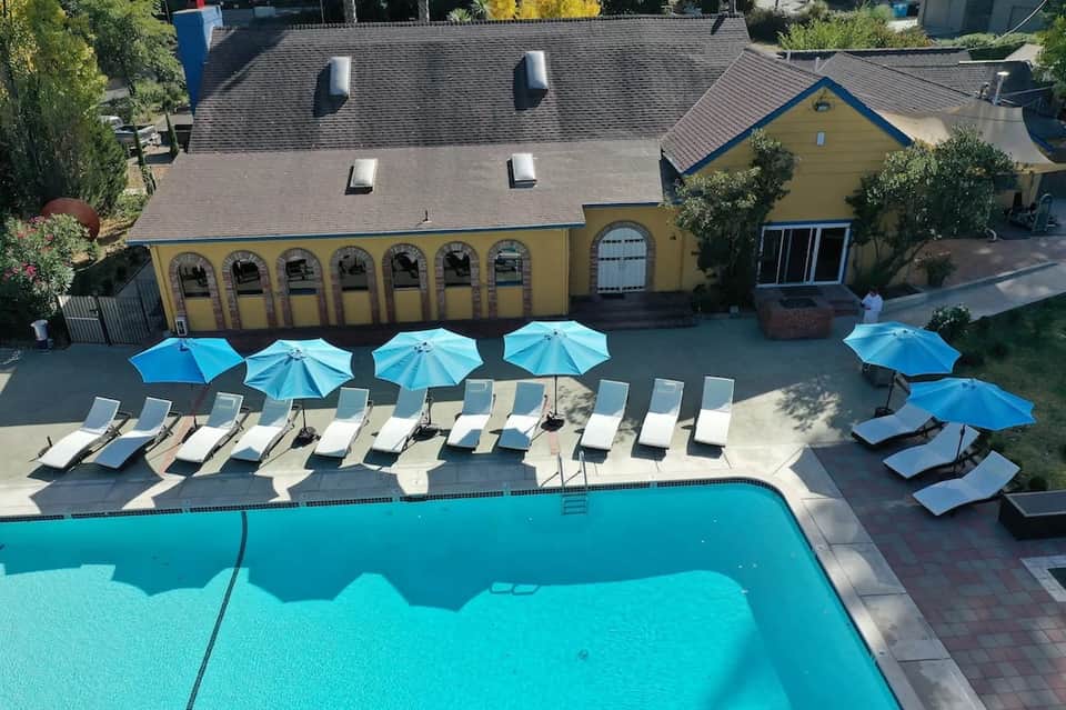Aerial view of resort pool with yellow building, blue umbrellas, white lounge chairs, and brick deck