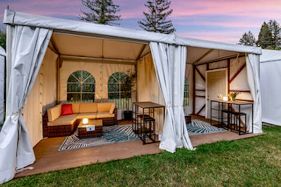 Outdoor glamping setup with white draped tent, orange sofa, and forest backdrop at dusk