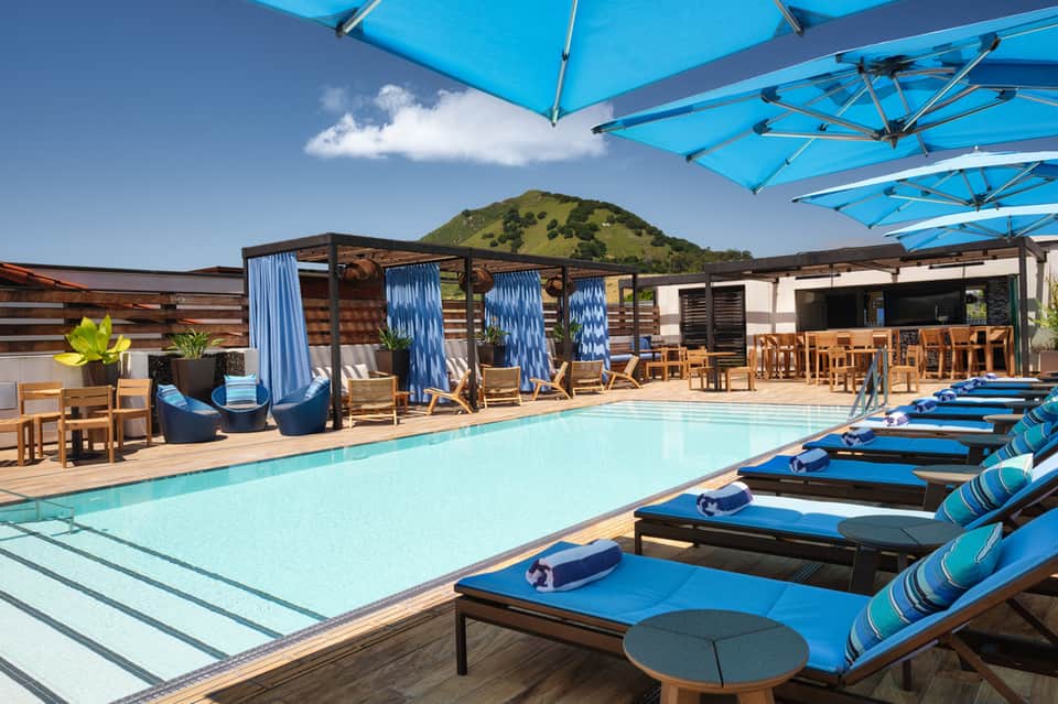 Resort pool deck with blue umbrellas, lounge chairs, wooden pergolas, and mountain views