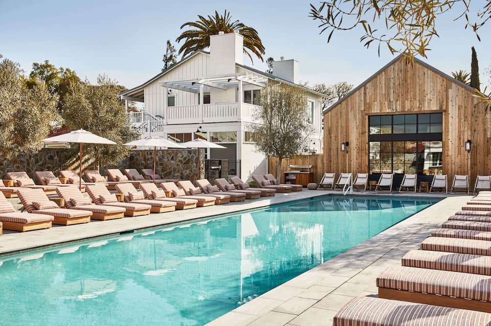 Resort pool area with loungers, umbrellas, modern wooden pavilion, and white building