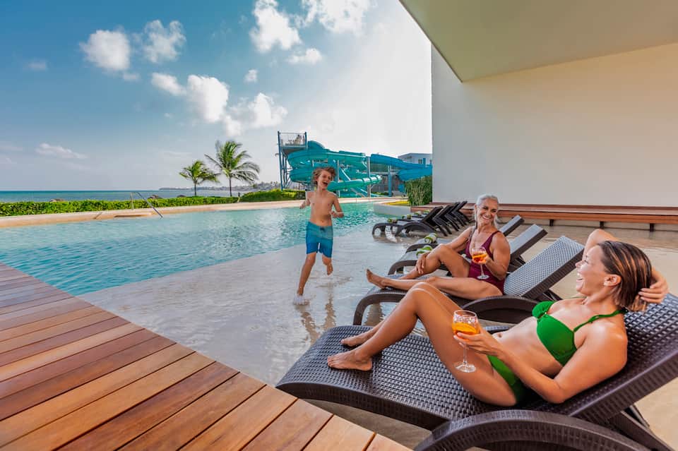 Beachfront resort pool with guests relaxing on lounge chairs and water slide in background